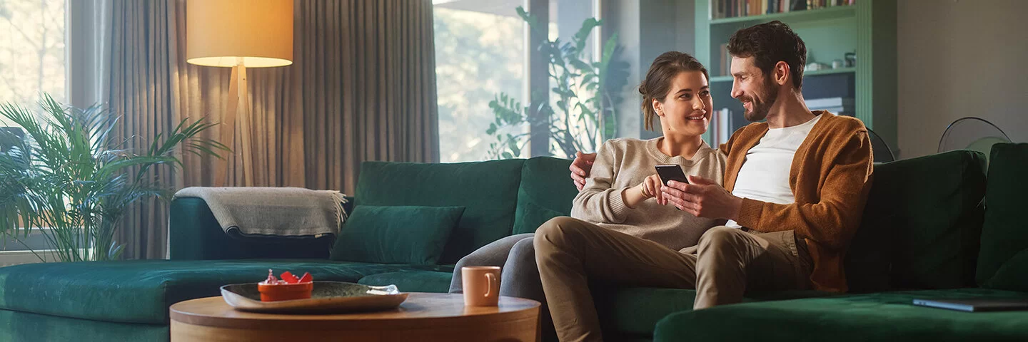Ein Pärchen in den Dreißigern sitzen nah beieinander auf dem Sofa und lächeln sich an. Der Mann hält ein Smartphone in seiner linken Hand, die Frau zeigt mit ihrer rechten Hand auf das Display.